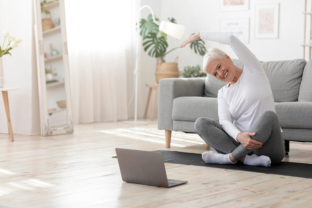 Senior woman sitting on floor in living room in front of couch stretching and looking at laptop screen