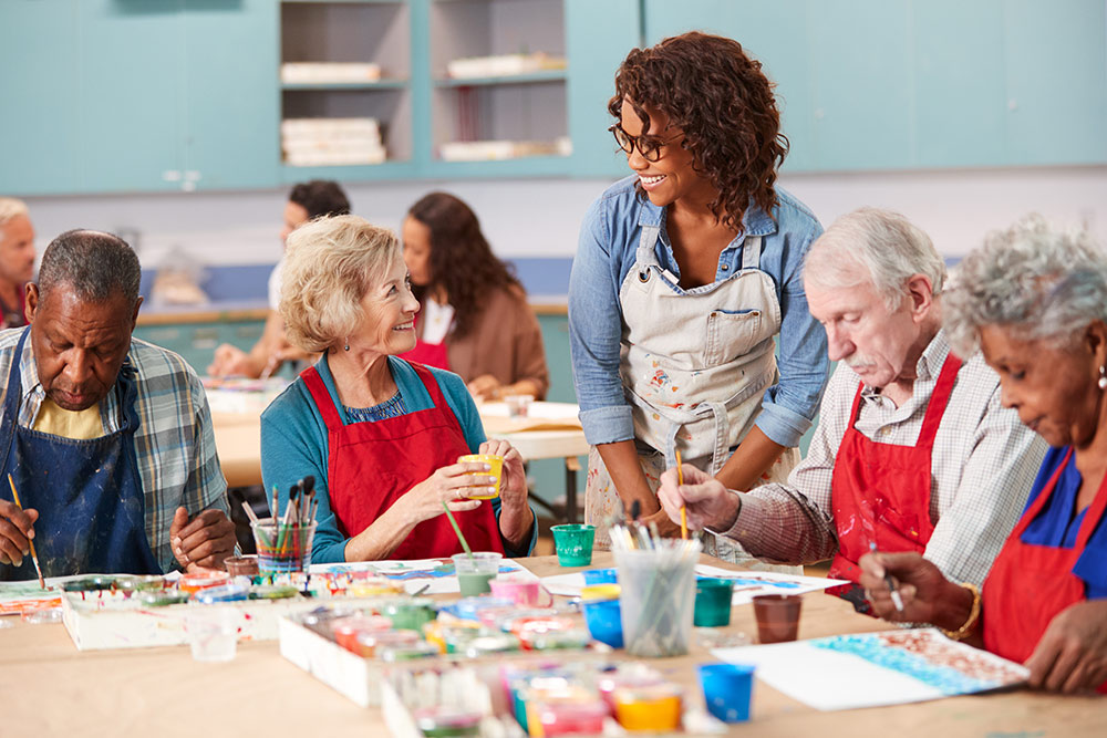 Senior art class with 4 seniors sitting around table painting and instructor watching over them all smiling