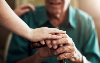 Care associate’s hand on an aging man’s hand holding a cane.